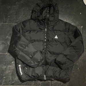 Svart pufferjacka från Nike ACG - Svart pufferjacka från Nike ACG med huva och dragkedja framtill. Jackan har Primaloft-isolering och Therma-FIT ADV-teknologi för extra värme. Snyggt ACG-logga på bröstet och Primaloft-detalj på ärmen. Perfekt för kalla dagar och har en clean, modern look.