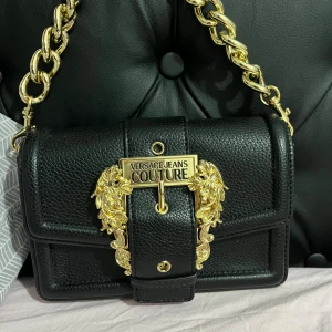 Svart axelväska från Versace Jeans Couture - Lyxig Versace Jeans Couture väska i toppskick, helt ny och aldrig använd. Köpt för 3 189 kr, säljes nu för 2 300 kr – ett riktigt kap! 💸  Väskan har fantastiska gulddetaljer, den ikoniska Versace-loggan och en elegant design som lyfter varje outfit. Den känns både sofistikerad och trendig, perfekt till både vardag och fest.  👜 Äkta vara – köpt i butik (kvittot tyvärr borttappat) 💛 Helt ny, inga repor eller märken 💫 Säljs endast för att jag inte använder den  📦 Skickas välpackad och spårbart dire