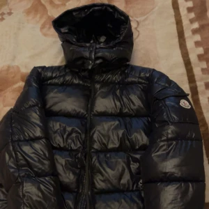 Svart Moncler pufferjacka med huva - Säljer en svart pufferjacka från Moncler med ikonisk logga på ärmen och coolt serietidningsfoder på insidan. Jackan har huva, dragkedja framtill och en riktigt schysst, glansig finish. Perfekt för dig som vill ha en stilren och varm jacka med streetvibe.