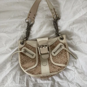 Beige axelväska från DKNY med kedja - Snygg beige axelväska från DKNY med mönstrat tyg och detaljer i skinn. Väskan har silverfärgade kedjor på axelremmen, två dragkedjor framtill och en stor spänndetalj. Jag fick den i en present men har aldrig använt🥰