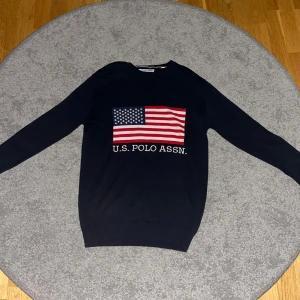 Mörkblå tröja U.S. Polo Assn. med USA flagga - Snygg mörkblå långärmad tröja från U.S. Polo Assn. med stort amerikanskt flaggtryck på bröstet och vit text under. Tröjan har rund hals och är gjord i mjukt bomullsmaterial. Perfekt för dig som gillar klassisk streetstyle. Oanvänd 