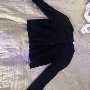 Svart kofta med knappar, stl S - En klassisk svart kofta med rund hals och knäppning framtill. Koftan har långa ärmar och enkla svarta knappar. Perfekt att slänga över en topp eller klänning för en clean look. Materialet känns mjukt och lätt, passar året runt.