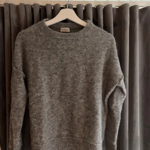 Grå stickad tröja By Malene Birger - Supermjuk och fluffig grå stickad tröja från By Malene Birger. Tröjan har rund halsringning, lång ärm och är tillverkad i en lyxig mix av alpacka, ull, polyamid och elastan. Perfekt för lager på lager under kyliga dagar.