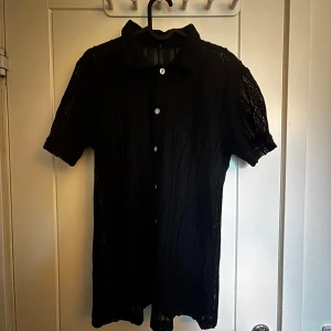 Svart kortklänning med knappar - Snygg svart kortklänning med krage och knappar framtill. Klänningen har korta ärmar och är tillverkad i ett stickat material med diskret struktur. Perfekt för dig som gillar enkel men stilren design med lite edge.