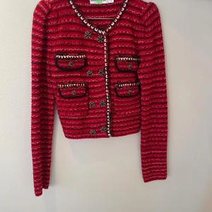 Röd cropped kofta från Self-Portrait - Säljer en röd cropped kofta från Self-Portrait med svarta och vita ränder, dekorativa knappar och fyra fickor framtill. Koftan är stickad och har lång ärm samt coola detaljer längs kanterna. Perfekt för dig som vill sticka ut med en trendig look.