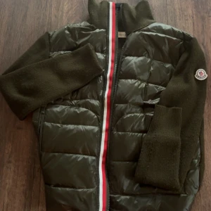 Mörkgrön pufferjacka från Moncler - Snygg mörkgrön pufferjacka från Moncler med stickade ärmar och krage. Jackan har quiltad front i glansigt material, dragkedja med rödvit rand och Moncler-logga på ärmen. Perfekt för dig som vill ha en cool och varm jacka med exklusiv känsla.