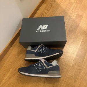  New Balance 574 - Snygga mörkblå sneakers från New Balance med klassisk N-logga på sidan. Skorna har grå och vita detaljer, rund tå och platt sula. Ovandelen är i mocka och mesh för både stil och komfort. Perfekt för dig som gillar streetwear och sportig look. Storlek 44 men passar 43