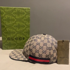 Gucci keps med GG-mönster och röd detalj - Snygg beige keps från Gucci med klassiskt GG-mönster över hela kepsen och en svart/röd rand baktill. Justerbar rem i bak och insida i svart bomull. Perfekt för dig som vill ha en lyxig och trendig accessoar.