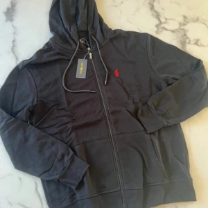 Svart hoodie från Polo Ralph Lauren XXL - Svart hoodie från Polo Ralph Lauren i storlek XXL. Tröjan har dragkedja, huva med snörning och en röd broderad logga på bröstet. Tillverkad i 100% bomull för skön och mjuk känsla. Perfekt för chill dagar och streetwear-stil.