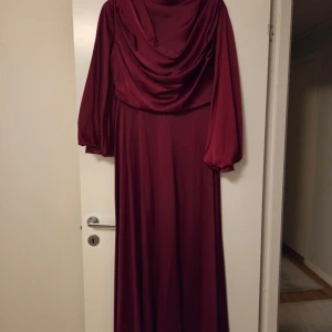 Vinröd långklänning med drapering - Elegant vinröd långklänning med mjukt fall och draperad framsida. Klänningen har långa, lätt puffade ärmar och en klassisk siluett som ger en lyxig känsla. Tillverkad i ett glansigt, följsamt material som ger ett exklusivt intryck.