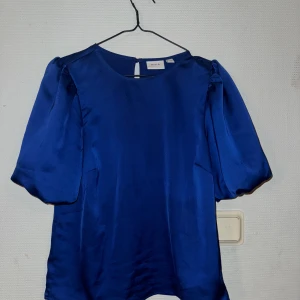 Blå satinblus från VILA - Säljer en snygg blå blus från VILA i glansigt satinmaterial. Blusen har puffiga korta ärmar och rund halsringning. Perfekt för dig som gillar stilrena och trendiga plagg med lite extra volym på axlarna. Färgen är djupblå och tyget känns lyxigt. Endast testad, prisen lappen ärborta! 