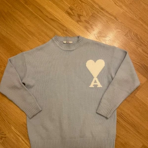Ljusblå stickad tröja från Ami - Snygg ljusblå stickad tröja från Ami med ett stort vitt hjärta och bokstaven A på bröstet. Tröjan har rund hals, ribbade muddar och långärmad passform. Perfekt för dig som gillar stilrena detaljer och vill ha något som sticker ut.