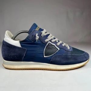 Blå sneakers från Philippe Model - Snygga blå sneakers från Philippe Model med detaljer i mocka och mesh, vita snören och vit häl. Skorna har en ikonisk sköld på sidan och en klassisk gummisula. Perfekta för dig som gillar en sportig och stilren look.