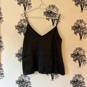 Svart linnetopp  - Snygg svart linnetopp lager 157 i storlek S. Toppen har smala axelband och en v-ringad halsringning. Materialet är mjukt och har en lätt glansig finish som ger en stilren look. Perfekt att matcha med jeans eller kjol för en cool vibe.