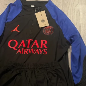 PSG tracksuit - Säljer en svart och blå långärmad PSG x Jordan fotbollströja med röd logga och tryck från Qatar Airways. Tröjan har dragkedja vid halsen och Paris Saint-Germain emblem på bröstet. Materialet är lätt och andas, perfekt för träning eller match. 10/10
