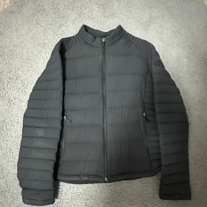 Svart pufferjacka Moose Knuckles XL - Svart pufferjacka från Moose Knuckles i storlek XL. Jackan har quiltad design, hög krage och två sidofickor med dragkedja. Materialet är syntet och polyester, perfekt för kalla dagar. Snygg logga på insidan och diskret emblem på ryggen.