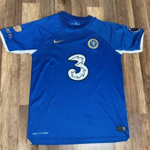 Chelsea FC blå Nike fotbollströja - Chelsea FC matchtröja i blått med guldiga detaljer och vit logga på bröstet. Kortärmad modell med broderat Chelsea-märke och Nike swoosh. Tillverkad i lätt, ventilerande polyester med vita och guldiga inslag på ärmarna. Perfekt för dig som vill representera ditt lag.