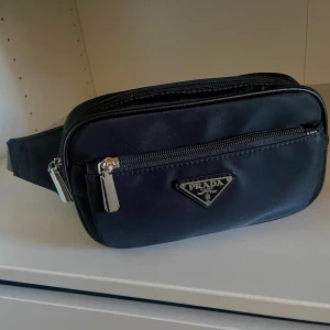 P midje väska i svart  - Snygg svart midjeväska från P med klassisk logga framtill. Väskan har två stora fack med dragkedja och justerbart band med spänne. Tillverkad i slitstark syntet med silverfärgade detaljer. Perfekt för att bära det viktigaste och hålla stilen. Inte andvänd 