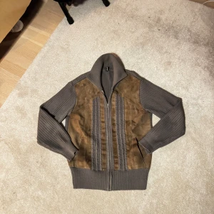 Brun och grå mocka-stickad cardigan - Snygg cardigan med brun mocka framtill och grå stickade partier på ärmar, sidor och krage. Jackan har dragkedja, ribbade muddar och kabelstickade detaljer längs framsidan. Perfekt för dig som gillar en unik och chill stil.