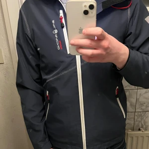 Mörkblå vindjacka från Sail Racing - Snygg mörkblå vindjacka från Sail Racing med vit dragkedja och röda detaljer. Jackan har huva, lång ärm och flera praktiska fickor med dragkedja. Insidan är röd mesh och jackan har coola vita tryck på ärmar och huva. Perfekt för dig som gillar sportig stil.