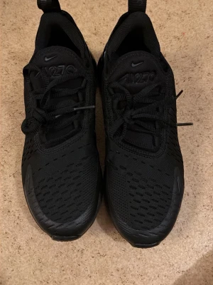 Svarta Nike Air Max 270 sneakers - Säljer ett par helsvarta Nike Air Max 270 sneakers med klassisk luftig mesh-ovandel och synlig Air-enhet i hälen. Skorna har snörning och en sportig, modern look som passar perfekt till streetwear. Swoosh-logga på sidan och diskreta detaljer.