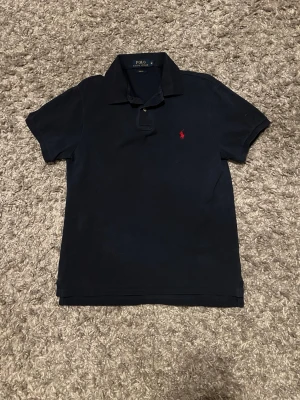 Marinblå pikétröja från Polo Ralph Lauren - Klassisk marinblå pikétröja från Polo Ralph Lauren med korta ärmar och krage. Tröjan har den ikoniska röda logotypen broderad på bröstet och är tillverkad i mjuk bomull. Perfekt för en stilren och avslappnad look.