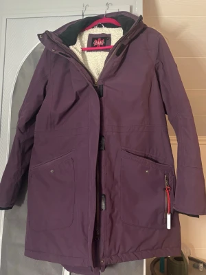 Lila parkasjacka  - Snygg lila parkasjacka från Ahkka med fuskpäls på huvan och mysigt teddyfoder. Jackan har dragkedja, stora fickor framtill och detaljer i svart. Perfekt för kalla dagar med sin långa passform och varma material.