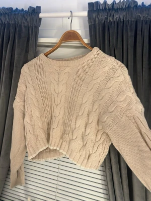 Beige croppad kabelstickad tröja - Säljer en beige croppad kabelstickad tröja med breda flätmönster och ribbade muddar. Tröjan har lång ärm och en loose fit som ger en avslappnad vibe. Perfekt till höst och vinter för en mysig och trendig look.