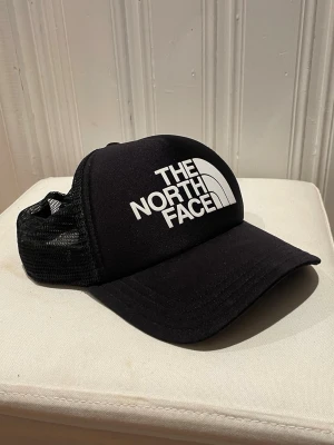 Svart keps från The North Face - Säljer en svart keps från The North Face med vit logga framtill. Kepsen har mesh på sidorna och bak för extra ventilation och en klassisk böjd skärm. Perfekt för dig som vill ha en stilren och sportig look.