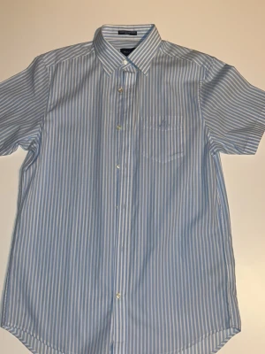 Blåvit randig kortärmad skjorta GANT - Säljer en fräsch blåvit randig skjorta från GANT i storlek S. Skjortan har korta ärmar, klassisk krage, bröstficka med broderad logga och knäppning framtill. Tillverkad i mjuk bomull som känns skön mot huden. Perfekt till jeans eller shorts.