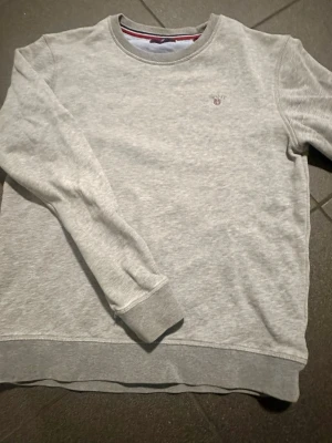 Grå sweatshirt från Gant - Klassisk ljusgrå sweatshirt från Gant med rund hals och diskret broderad logga på bröstet. Tröjan har ribbade muddar vid ärmslut och nederkant, perfekt för en clean och enkel stil. Finns en fläck på bröstet vänster sida. Syns om man zoomar bilden 