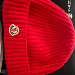 Röd Moncler mössa - Säljer en snygg röd ribbstickad mössa från Moncler med klassisk logga framtill. Mössan är i ett mjukt och varmt material som passar perfekt till kyliga dagar. Enkel och stilren design som lyfter vilken outfit som helst. Äkta med kvitto