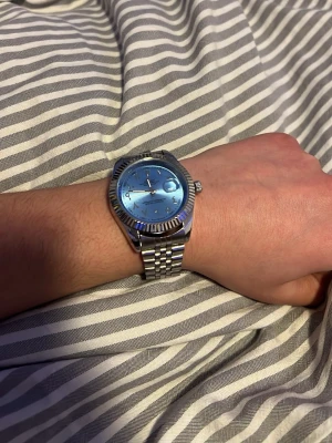 Rolle klocka  - Bilderna visar en silverfärgad klocka med blå urtavla och metallänk, men ingen väst syns.