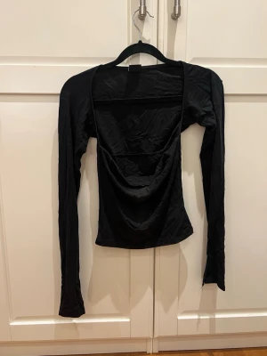 Svart långärmad topp  - Säljer en svart långärmad topp med snygg drapering framtill och djup urringning. Toppen har en tight passform och är tillverkad i ett mjukt, stretchigt material som sitter skönt på kroppen. Perfekt att styla med jeans eller kjol.