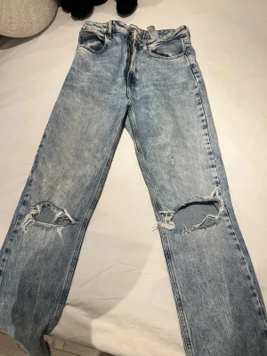 Ljusblå slitna jeans med hål - Säljer ett par ljusblå jeans med stora slitningar och hål vid knäna. Klassisk femficksmodell med rak passform och hög midja. Jeansen har en vintage tvätt och är tillverkade i bomull för en skön känsla.