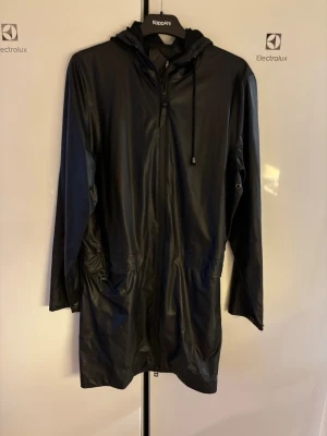 Svart regnjacka med huva och fickor - Svart regnjacka med huva och dragsko, tillverkad i syntetmaterial. Jackan har lång ärm, två stora fickor framtill och en half zip-dragkedja. Perfekt för regniga dagar och enkel att matcha med olika outfits.