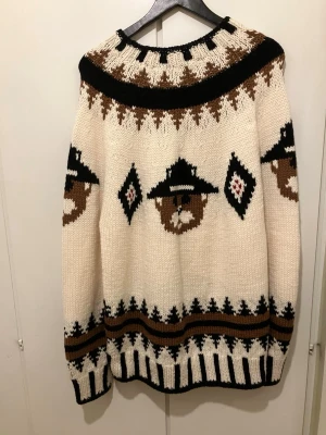 Stickad tröja med björnmönster - Unik stickad tröja i vitt med brunt, svart och beige mönster. Tröjan har björnar med hattar runtom, grafiska detaljer vid hals, ärmslut och nederkant. Oversized passform och rund halsringning. Perfekt för dig som gillar statement-plagg och retrovibbar.