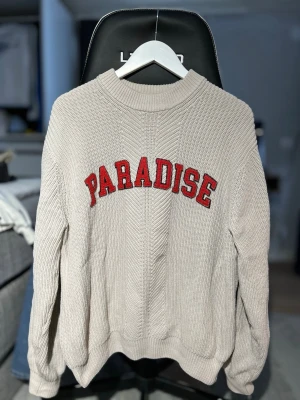 Beige stickad tröja med text från Vailent - Säljer en beige stickad tröja med röd text 'PARADISE' broderad över bröstet. Tröjan har rund halsringning, ribbade muddar och en relaxed fit. Perfekt för dig som gillar en enkel men ändå statement look.