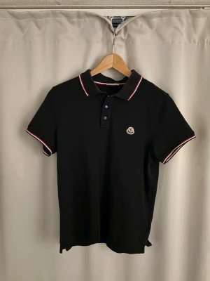 Svart Moncler pikétröja med röd/vit kant - Klassisk mörkblå pikétröja från Moncler med röd och vit kant på krage och ärmslut. Tröjan har knappslå framtill och Moncler-logga broderad på bröstet. Perfekt för en clean och stilren look. mörkblå 