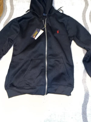 Svart hoodie från Polo Ralph Lauren - Svart hoodie med dragkedja från Polo Ralph Lauren. Klassisk design med huva, två fickor framtill och röd broderad logga på bröstet. Tillverkad i mjukt material som är skönt att bära. Perfekt för en avslappnad och stilren look.