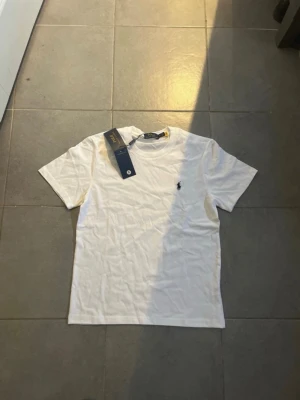 Vit t-shirt från Polo Ralph Lauren - Klassisk vit t-shirt från Polo Ralph Lauren med rund halsringning och ikoniska broderade loggan på bröstet. T-shirten är i bomull och har en normal passform med korta ärmar. Perfekt basplagg för en stilren look.