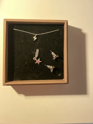Smyckesset med blixt,stjätna och måne - Säljer ett silvrigt smyckesset med halsband med blixtberlock, armband med sjärnberlock och ett par örhängen formade som små månar. Perfekt för dig som gillar rymden eller bara vill ha lite matchande smycken. Materialet är metall i silverfärg.Kan köpa en och en 25kr styck