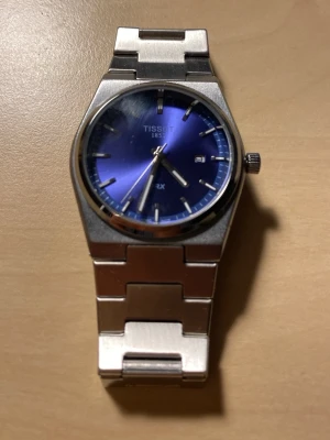 Tissot PRX klocka med blå urtavla 40mm - Snygg Tissot PRX klocka i rostfritt stål med länkarmband och blå urtavla. Klockan har minimalistiska index, datumfönster och klassisk rund boett. Perfekt accessoar för dig som gillar stilren design och kvalitetskänsla.se bilder för egen uppfattning av skick. Obs lite repor på backsidan.