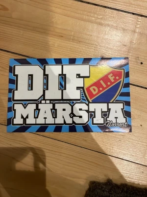 DIF Märsta klistermärke - Klistermärke med texten 'DIF Märsta' och DIF:s klassiska sköld i blått, gult och rött. Bakgrunden har blå strålar och texten är vit med svart kant. Perfekt för att visa sitt stöd för Djurgården och Märsta.