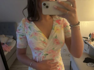 Blommig omlottklänning med puffärm - Säljer en lång, blommig omlottklänning med puffärmar och v-ringning. Klänningen har ett vackert pastellmönster i rosa, gul och grön på vit botten. Tillverkad i ett lätt och luftigt material, perfekt för sommarens alla tillfällen.