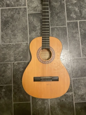 Klassisk akustisk gitarr Valencia - Säljer en klassisk akustisk gitarr från Valencia med naturfärgad träkropp och mörkbrunt greppbräde. Gitarren har sex nylonsträngar, dekorativ rosett runt ljudhålet och silverfärgade stämskruvar. Perfekt för dig som vill börja spela eller behöver ett extra instrument.