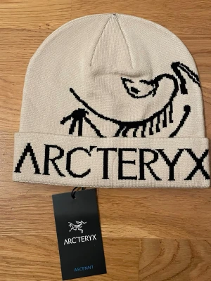 Arc’teryx Ascent Beanie – Beige – Oanvänd!! - Snygg beige mössa från Arc'teryx med svart logotyp och grafiskt mönster framtill. Mössan är ribbstickad och har uppvikt kant med tydlig branding. Perfekt för dig som gillar stilrena accessoarer med outdoor-vibe.