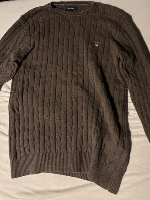 Brun kabelstickad tröja från GANT - Säljer en brun kabelstickad tröja från GANT med rund halsringning och diskret logga broderad på bröstet. Tröjan har ribbade muddar vid ärmslut och nederkant, vilket ger en klassisk och stilren look. Perfekt för lager på lager under kyliga dagar. Nypris cirka 1300