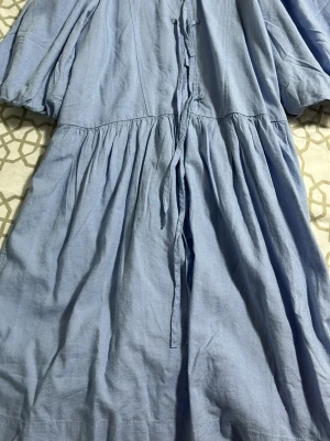 Ljusblå omlottklänning i linne - Säljer en ljusblå omlottklänning med knytband i midjan och puffiga ärmar. Klänningen har en avslappnad passform och är gjord i ett luftigt linnematerial, perfekt för varma dagar. Enkel men stilren design som känns både trendig och bekväm.