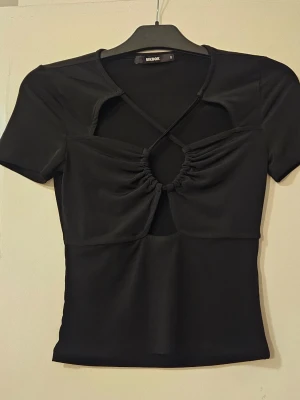 Svart topp med cut out från Bik Bok - Trendig svart topp från Bik Bok med korta ärmar, cut out-detaljer och snygg rynkning framtill. Toppen har en croppad passform och dekorativa band över bröstet som ger en cool look.